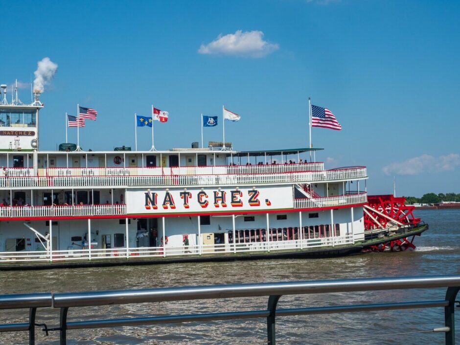 El Barco de Vapor Natchez con rueda de paletas navega en el río, ondeando banderas estadounidenses y estatales bajo un cielo azul