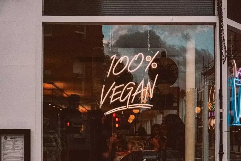 Neón con el texto '100% VEGAN' destaca en la ventana de un restaurante, invitando a los transeúntes a explorar opciones alimenticias compasivas y sostenibles