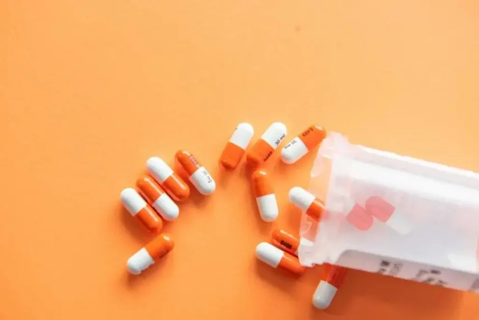 Cápsulas naranjas y blancas con inscripciones médicas esparcidas de un frasco de prescripción sobre fondo naranja