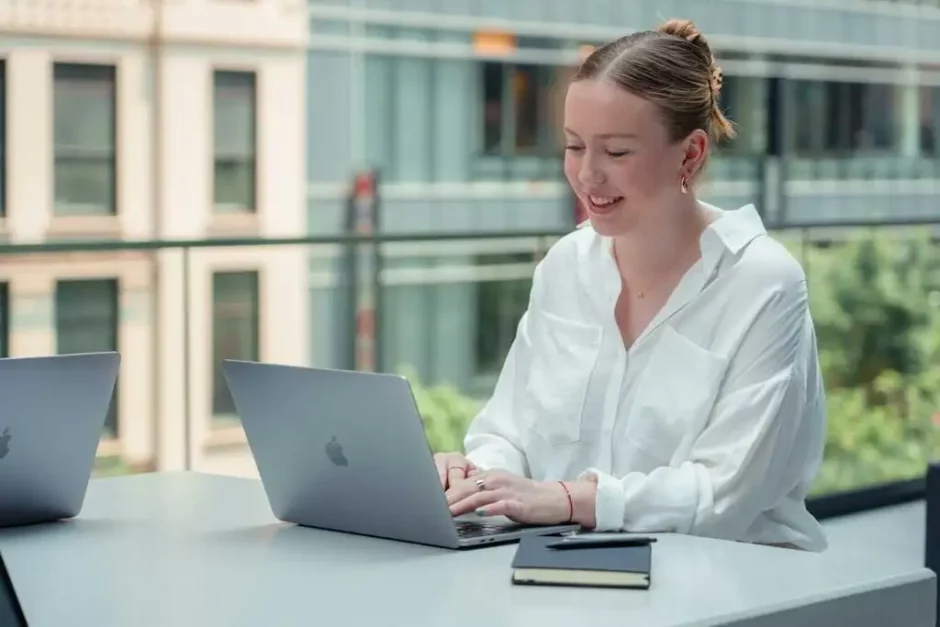 Una joven sonriente trabajando en dos laptops al aire libre en un espacio urbano moderno, representando el estilo de vida de un trabajador remoto o nómada digital