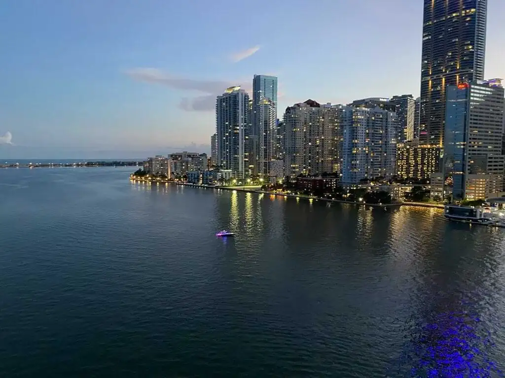 Vista aérea del anochecer en Miami, mostrando rascacielos iluminados reflejándose en la tranquila bahía, con luces de embarcaciones navegando suavemente
