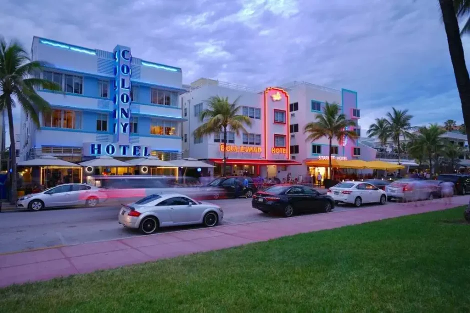 Vista crepuscular de Ocean Drive con hoteles Art Decó iluminados en neón, coches en movimiento y palmeras en Miami Beach, evocando una vibrante atmósfera nocturna