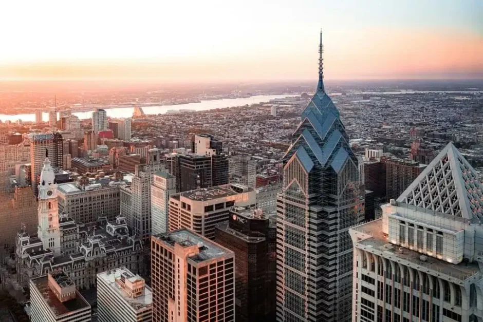 Vista aérea cautivadora del skyline de Filadelfia durante el atardecer. Edificios prominentes incluyen el ornamentado Ayuntamiento de Filadelfia con su icónica torre de reloj y rascacielos modernos, especialmente Liberty Place con su cima en forma de pirámide. La ciudad se extiende hacia el horizonte, y un río brilla a lo lejos