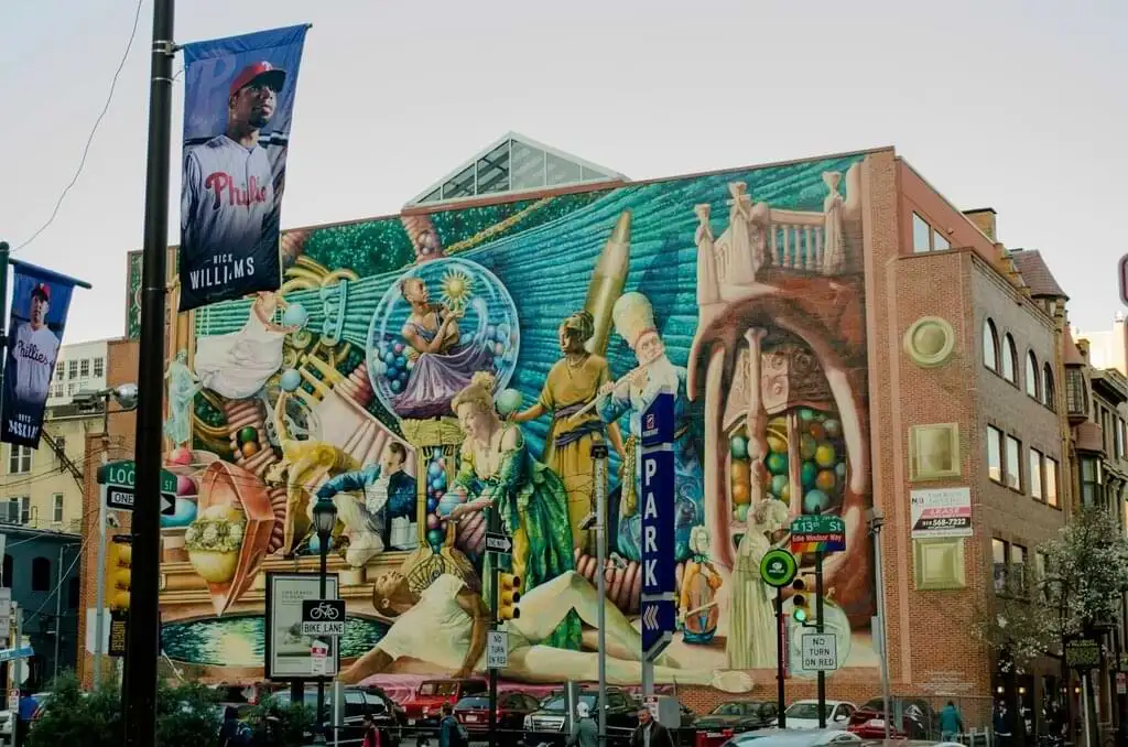 Un mural vibrante adorna la fachada de un edificio en Filadelfia. La obra de arte muestra una variedad de figuras coloridas en poses dinámicas, rodeadas de objetos y símbolos abstractos. La escena está ubicada en una esquina de la ciudad, con postes de señalización y pancartas de béisbol en primer plano