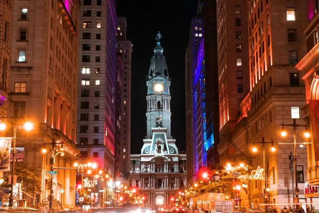 Vista nocturna de la ciudad de Filadelfia, enfocada en una calle concurrida flanqueada por edificios altos y luminosos. En el fondo, el Ayuntamiento de Filadelfia resplandece con su distintiva torre de reloj, enmarcada por las luces y el movimiento de la ciudad