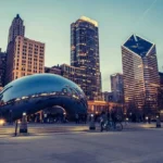 Fotografía de un anochecer en Chicago que muestra el icónico Cloud Gate, conocido como el Bean, reflejando los edificios iluminados del centro de la ciudad. En primer plano, personas pasean y disfrutan del ambiente urbano, mientras los rascacielos se alzan en el fondo bajo un cielo azul oscuro